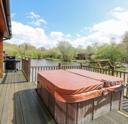 A deck with a covered hot tub and a barbecue overlooking a pond with trees and a wooden cabin at Woodpecker Lodge in Badwell Ash