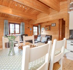 A wooden interior room with a dining table with chairs a vase with flowers leather sofas windows and kitchen appliances at Woodpecker Lodge in Badwell Ash