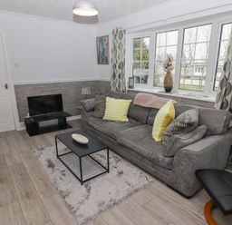 A living room with a gray sofa yellow and patterned cushions a black coffee table a rug a small television and a window with patterned curtains at 48 Gower Holiday Village in Scurlage