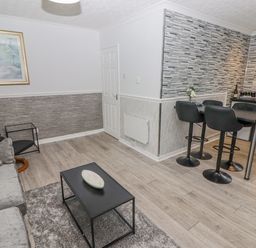 A living area with a gray sofa a black coffee table a small side table and a kitchen area with black stools and a tiled wall at 48 Gower Holiday Village in Scurlage