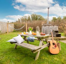An outdoor garden area with a wooden picnic table set with food and drinks a guitar resting against the table a hammock and a wooden hot tub in the background at Wheal Jewel Lodge in Wendron near Helston