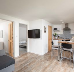 A kitchen and living area with wooden flooring a dining table with chairs a wall-mounted tv and bedrooms visible through doorways at Wheal Jewel Lodge in Wendron near Helston