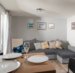 A dining table set with plates and glasses next to a grey sectional sofa with cushions and framed pictures on the wall at Wheal Prosper Lodge in Wendron near Helston