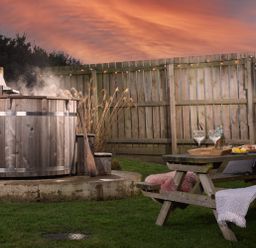 An outdoor area with a wooden hot tub emitting steam a wooden picnic table with drinks and food on it and a wooden fence in the background at Wheal Rose Lodge Wendron near Helston
