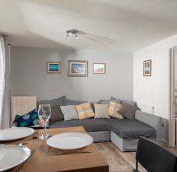 A living room with a grey sectional sofa and cushions a wooden dining table set with white plates and glasses and a doorway showing a bedroom with bunk beds at Wheal Rose Lodge in Wendron near Helston