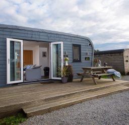 A wooden deck with picnic table and plants outside a small blue cabin with open doors at Wheal Tor Lodge Wendron near Helston