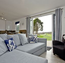 A living area with a light gray sectional sofa with decorative pillows and a dark armchair next to large sliding glass doors overlooking a garden at Wheal Tor Lodge Wendron near Helston
