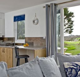 A kitchen corner with a wooden cabinet and countertop with a black chair and a plate of fruit next to a window and glass door with curtains and a gray sofa with cushions in the foreground at Wheal Tor Lodge in Wendron near Helston
