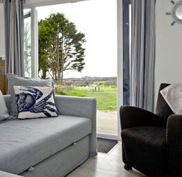 A living room with a gray sofa and crab cushion next to a dark armchair with a pink cloth beside a sliding glass door showing an outdoor view at Wheal Tor Lodge in Wendron near Helston