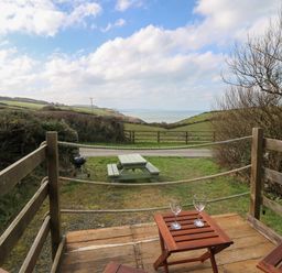 A wooden deck with two chairs and a table with two glasses overlooking a picnic table and green fields with a view of the sea at Sea the Stars in New Quay