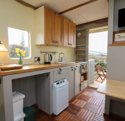 A small kitchen with wooden cabinets a sink a kettle a lamp and flowers on the counter with an open door leading to an outdoor seating area at Sea the Stars in New Quay