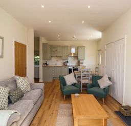 An open plan living area with a gray sofa and patterned cushions two green armchairs a wooden coffee table dining table with chairs and a kitchen with green cabinets at 2 Twizel Station Cottage Cornhill-on-Tweed near Norham