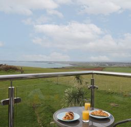 A balcony with a table holding two plates with croissants and two glasses of orange juice overlooking green fields and a coastline at 5 Ocean Reach in Bolberry
