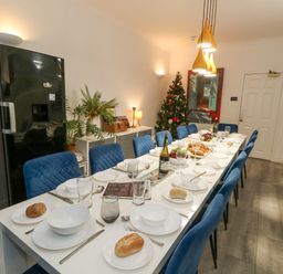 A dining room with a long table set with plates glasses and bread rolls blue chairs around the table a decorated Christmas tree near a door a black refrigerator and plants at Beachaven in Weymouth