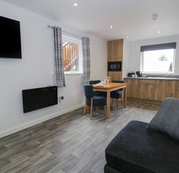 A kitchen and dining area with wooden cabinets a wooden table and blue chairs a gray sofa and a wall-mounted television at Classic Lodge 25 in Dyserth