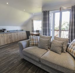 A living area with a sofa and cushions next to a window and open door with curtains and a kitchen area with cabinets and appliances at Premier Lodge 41 in Dyserth