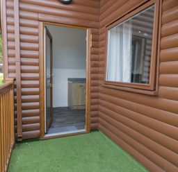 A balcony with artificial grass and wooden railing outside a lodge with an open door showing a kitchenette at Classic Lodge 15 in Dyserth
