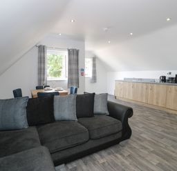 An open living area with a grey sectional sofa, dining table with four chairs by windows, and a kitchenette with wooden cabinets at Classic Lodge 15 in Dyserth