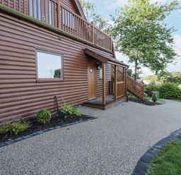 A brown wooden lodge with a small porch and stairs surrounded by greenery and a paved driveway at Luxury Lodge 46 in Dyserth