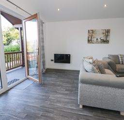 A living room with an open door to a balcony a grey sofa multiple cushions a wall-mounted electric fireplace and a painting at Luxury Lodge 53 in Dyserth