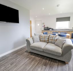 A living room with a grey sofa with cushions a wall-mounted television wooden dining table with blue chairs and a kitchenette at Luxury Lodge 62 in Dyserth