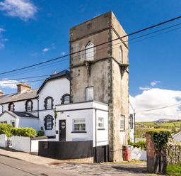 A street view of a tall stone tower attached to a white house with black trim and nearby greenery at The Old Coastguard Tower Buncrana Buncrana County Donegal