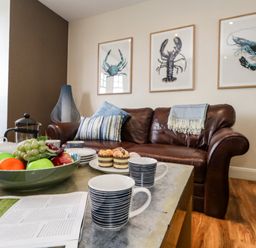 A living room with a brown leather sofa three framed sea creature pictures and a coffee table with mugs fruit and pastries at The Lobster Pot in Weymouth