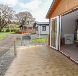 A wooden deck outside a house with an open door showing a living room with curtains and furniture a pathway a grassy area and two other similar houses in the background at Lodge 4 in St Asaph