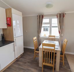 A dining area with a wooden table and four chairs next to a window with curtains in a kitchen space at Lodge 4 in St Asaph