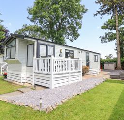 A white mobile home with a small deck and outdoor seating on a grass lawn surrounded by trees at Woodlands Lodge in Bontuchel near Ruthin