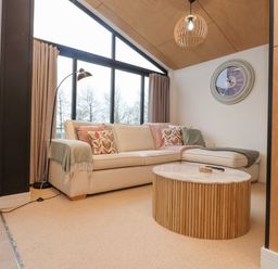A living room with a beige sectional sofa near large windows and a round wooden coffee table at Encil Gyfrinfa in Gwynus near Llithfaen