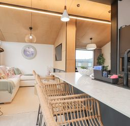 An open kitchen and living area with a countertop, rattan bar stools, a sofa, a wall clock, and a television at Encil Gyfrinfa in Gwynus near Llithfaen