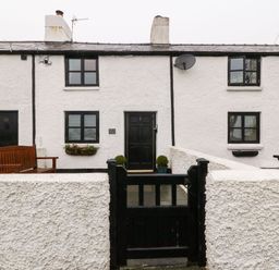 The front of a white cottage with a black door a black gate and a wooden bench at White Lilac Cottage in Brynsiencyn
