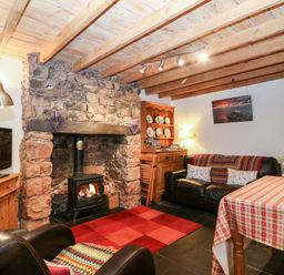 A living room with a stone fireplace black wood stove two black leather chairs a red checkered rug and a table with a checkered tablecloth at White Lilac Cottage in Brynsiencyn