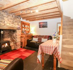 A living room with a stone fireplace a wood burning stove a red checkered tablecloth on a table and carpeted stairs at White Lilac Cottage Brynsiencyn