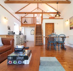 A living area with a brown leather sofa, a wooden coffee table with games on it, a dining table with blue chairs, and a wooden staircase leading to a loft at Joe's Barn in Mullion