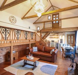 An open living room with brown leather sofas a wooden coffee table a rug and a kitchen with dining table and chairs at Berts Barn in Mullion