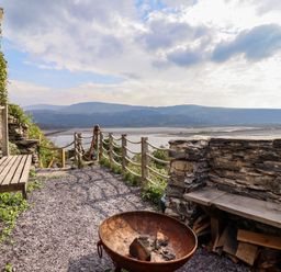 An outdoor seating area with wooden benches a fire pit stone walls and a view of hills and water at Pengarreg in Barmouth
