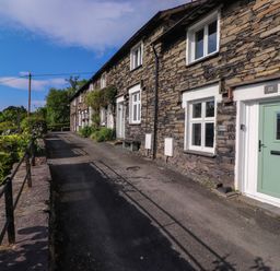 A row of stone cottages with white framed windows and a green door along a paved path with plants and a metal railing at Lavender Cottage in Coniston