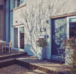 An exterior view of a house with a blue front door a glass sliding door a wooden deck with table and chairs and potted plants at Craster Reach in Craster