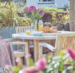 An outdoor wooden dining table with wooden chairs set with bread juice and flowers at Craster Reach in Craster