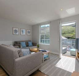 A living room with two sofas a coffee table and a TV on a stand with open door to a balcony showing a hillside outside at Rebecca in Mullion Cove near Mullion