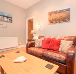 A living room with a brown leather sofa with cushions a wooden coffee table with a heart-shaped dish a lamp on a side table and landscape photos on the walls at Myrtle Cottage in Windermere