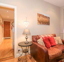 A living room with a leather sofa and red cushions a side table with a lamp and a hallway with wooden doors at Myrtle Cottage in Windermere