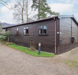 A dark brown wooden cabin with windows and a small garden area on a gravel path at Hayeswater Lodge in Burnside Park near Keswick