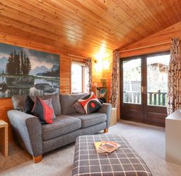 A living room with a gray sofa and patterned cushions an ottoman a TV on a cabinet and a large landscape picture on a wooden paneled wall at Hayeswater Lodge Burnside Park near Keswick