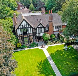 A large house with a tiled roof and black and white exterior surrounded by trees and a lawn with a stone path and outdoor chairs at Tudor Grange in Bournemouth