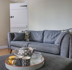 A grey sofa with patterned cushions and throw in a living room with a tray containing a teapot and mugs on a grey ottoman at 8 Biddlecombe Orchard in Bridport