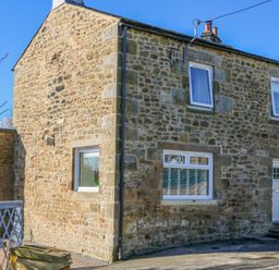 A stone house with white framed windows and a white door on a paved street at Bridge End Cottage in Wark