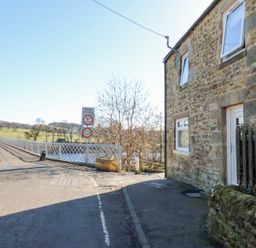 A narrow road leading to a metal bridge next to a stone house with windows and a door at Bridge End Cottage in Wark